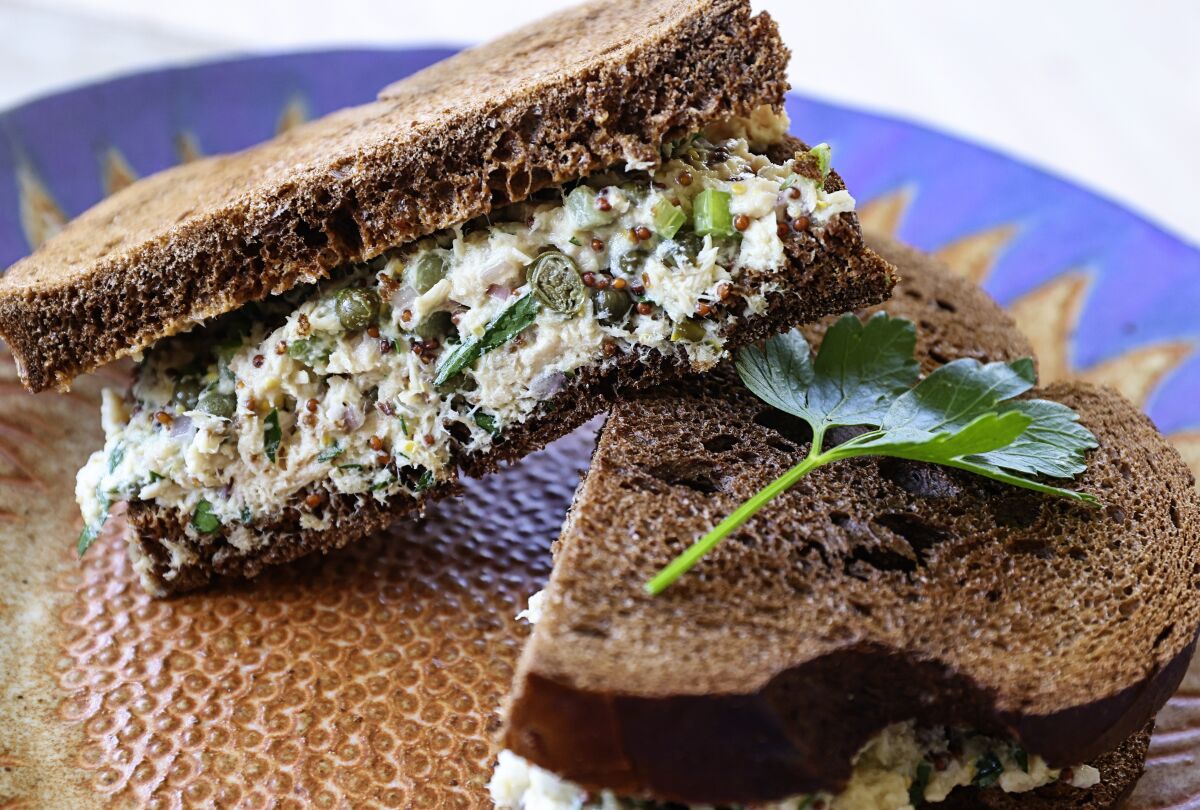 Halves of a Tuna, Caper and Cornichon Sandwich on Pumpernickel are stacked on a plate.