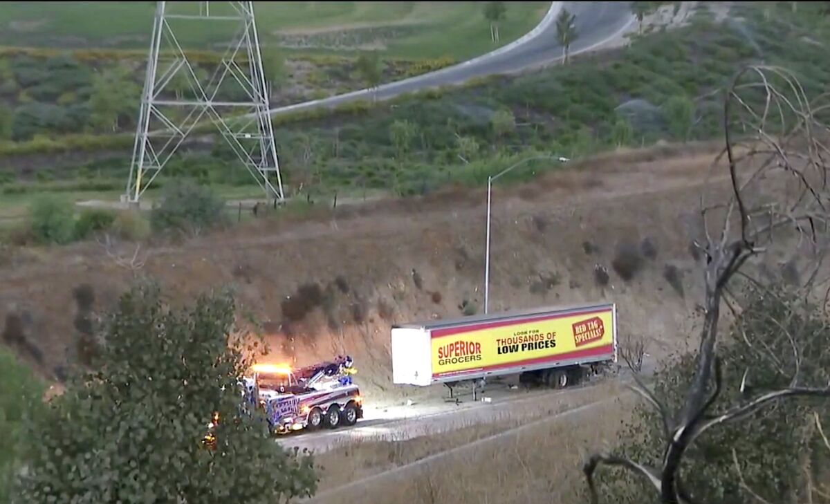 A tow truck waits in front of a big rig's load on a road surrounded by grass and trees.