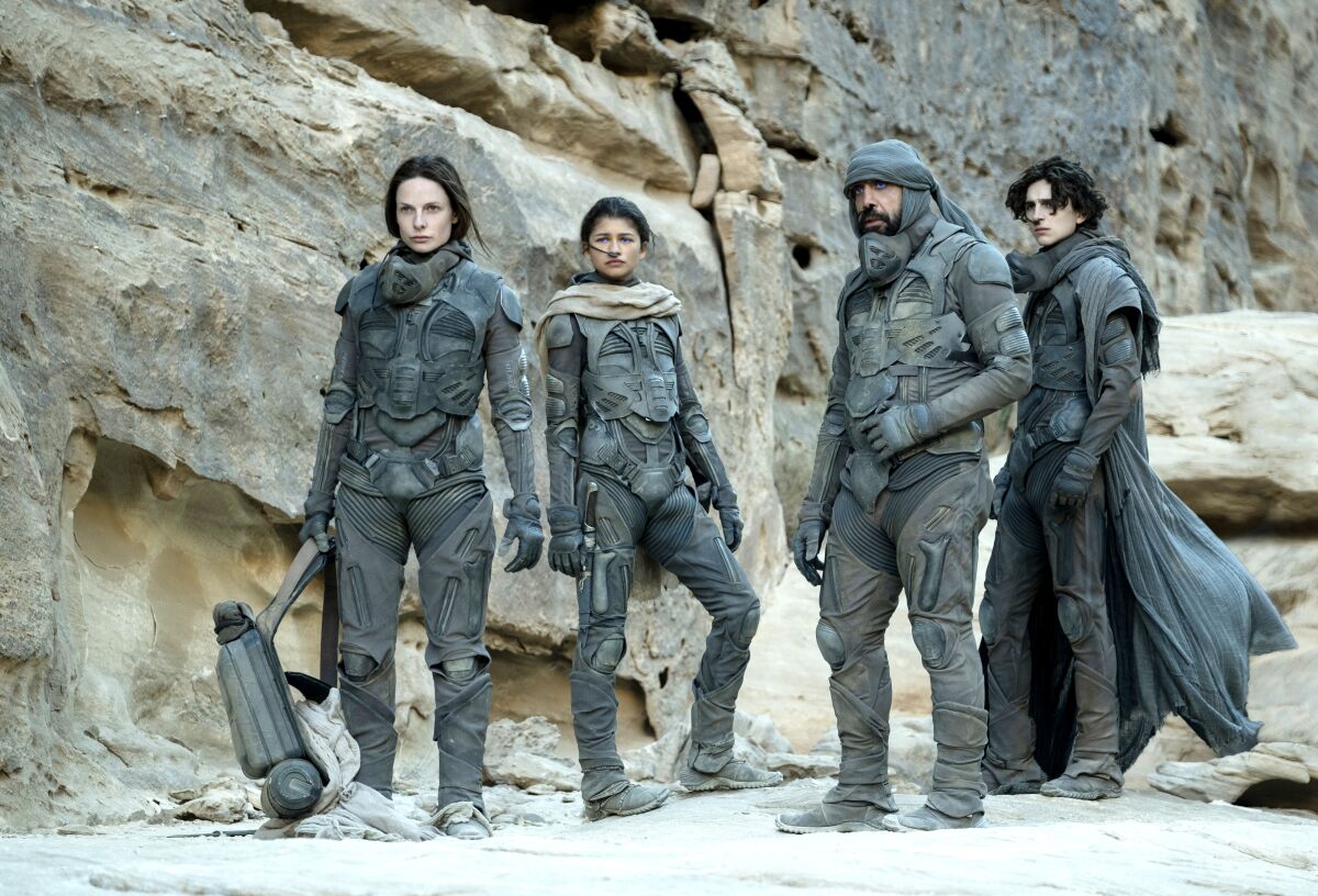 Four people in protective suits stand in a desolate landscape.