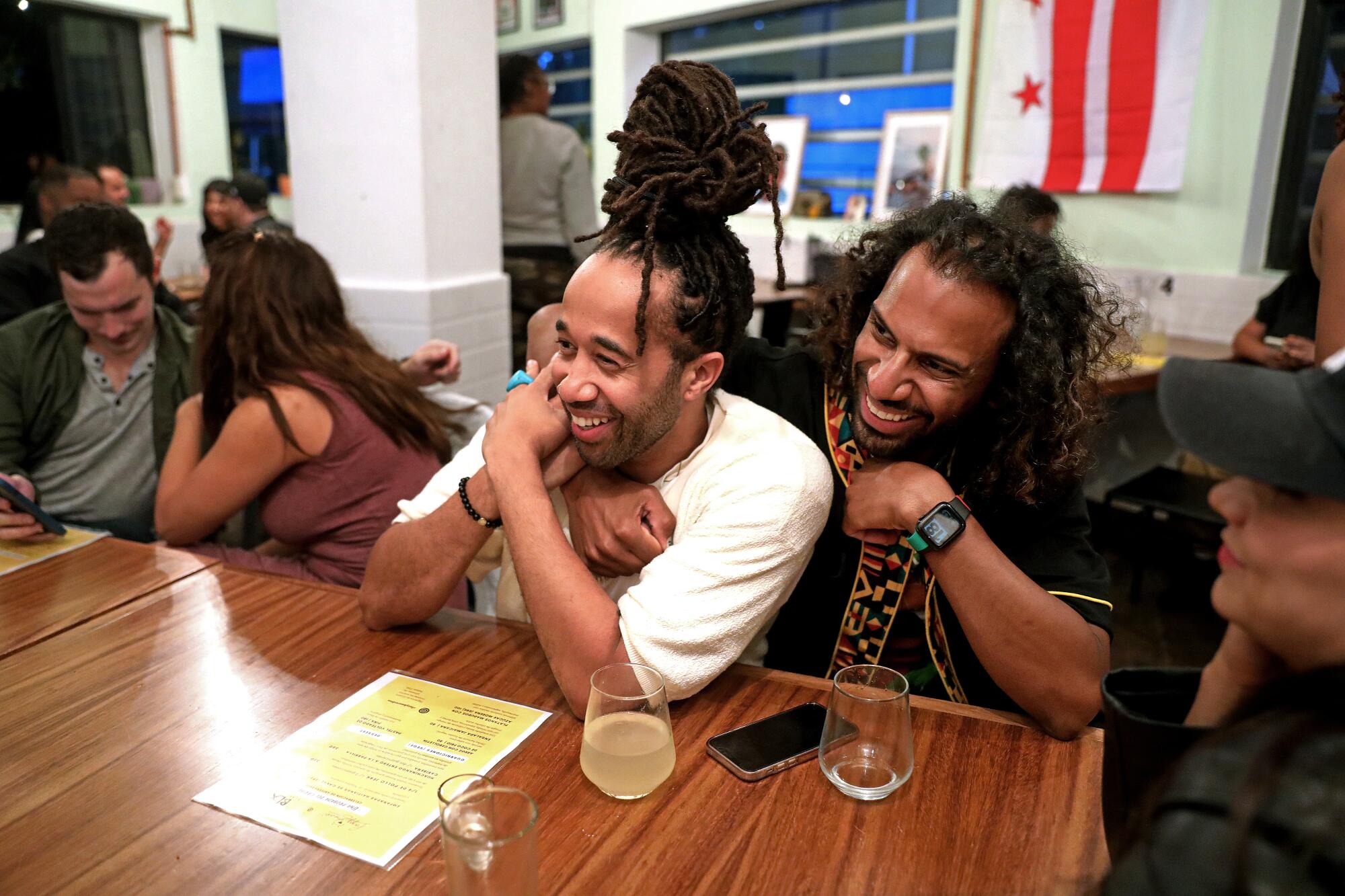 Two Black men smile and sit together at a restaurant table.