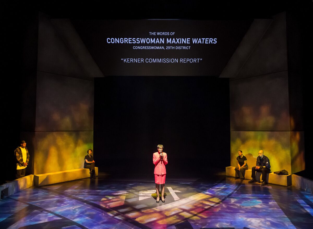 A balcony view of a stage with a woman standing center stage and other actors in the background