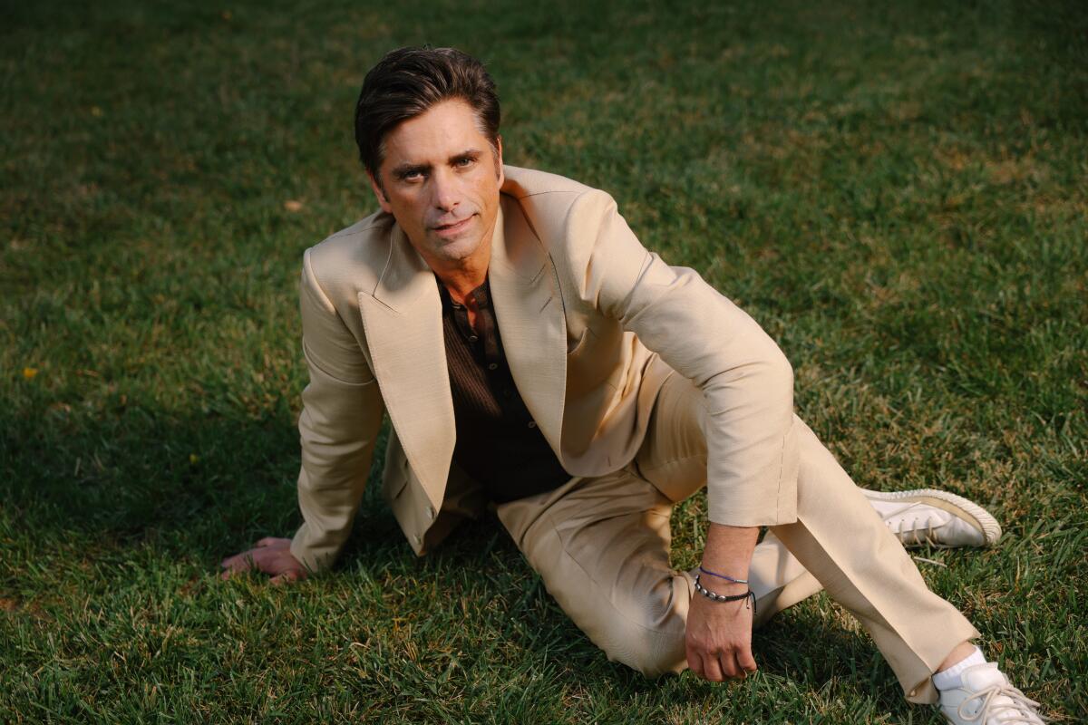 John Stamos smiles as he wears a suit and sits in the grass