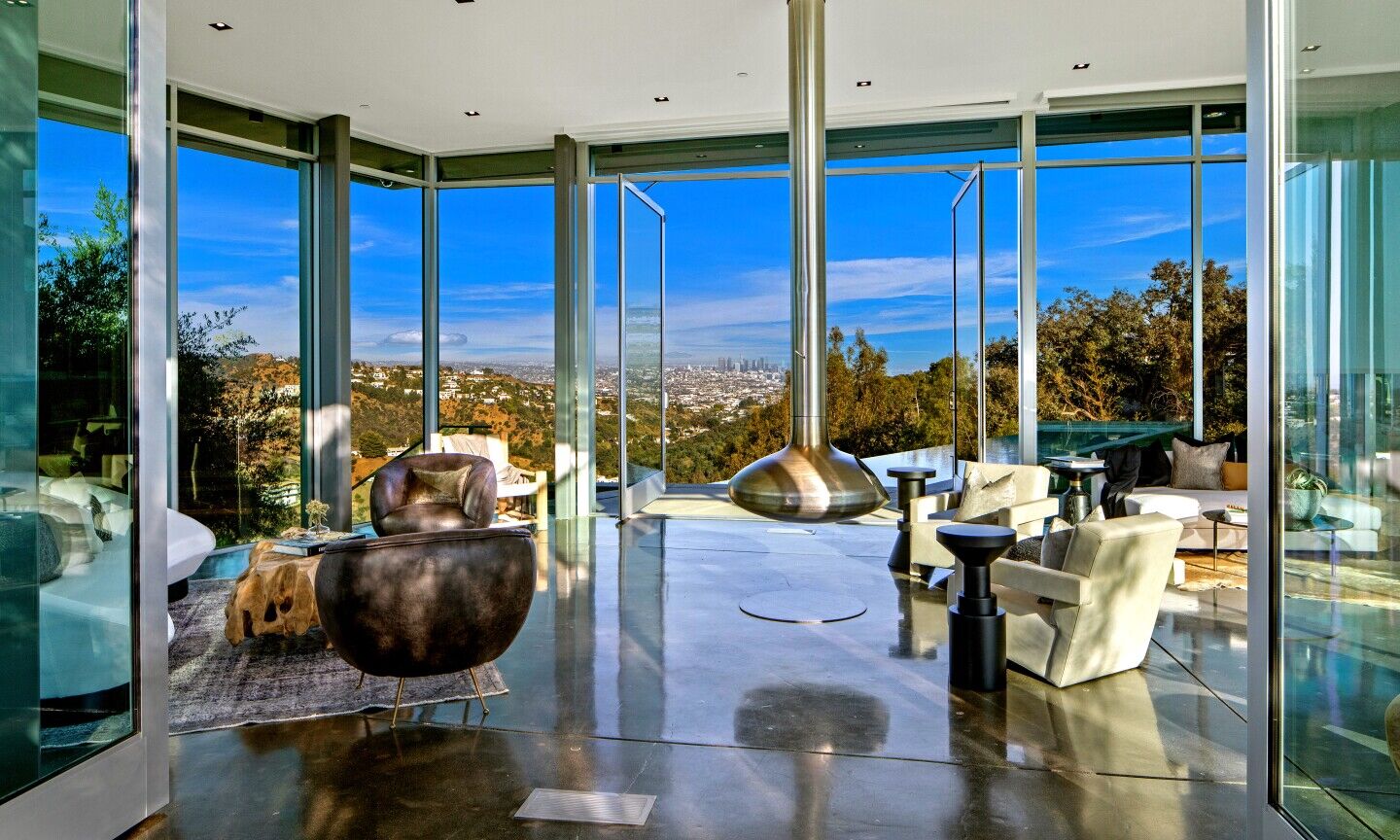 The living room has glass walls, a shiny floor and furniture overlooking greenery and sky.