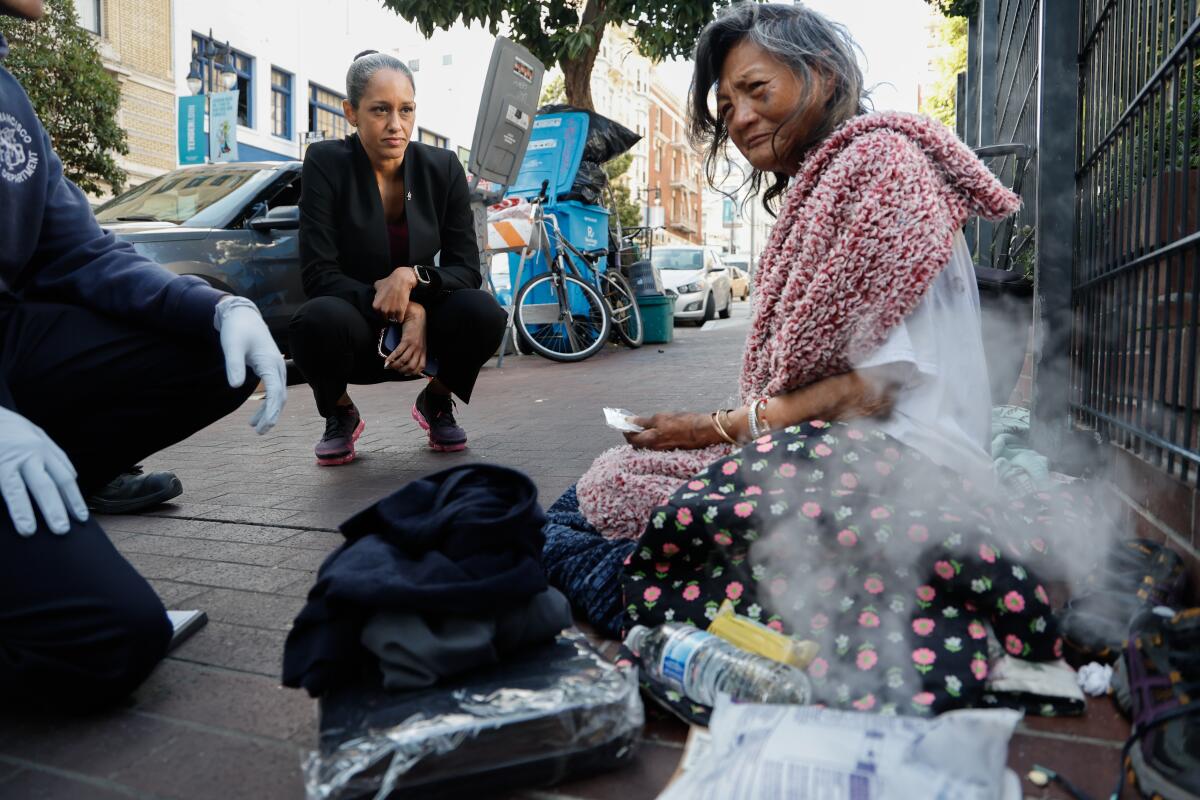 A woman squats on the sidewalk while talking to a homeless woman.