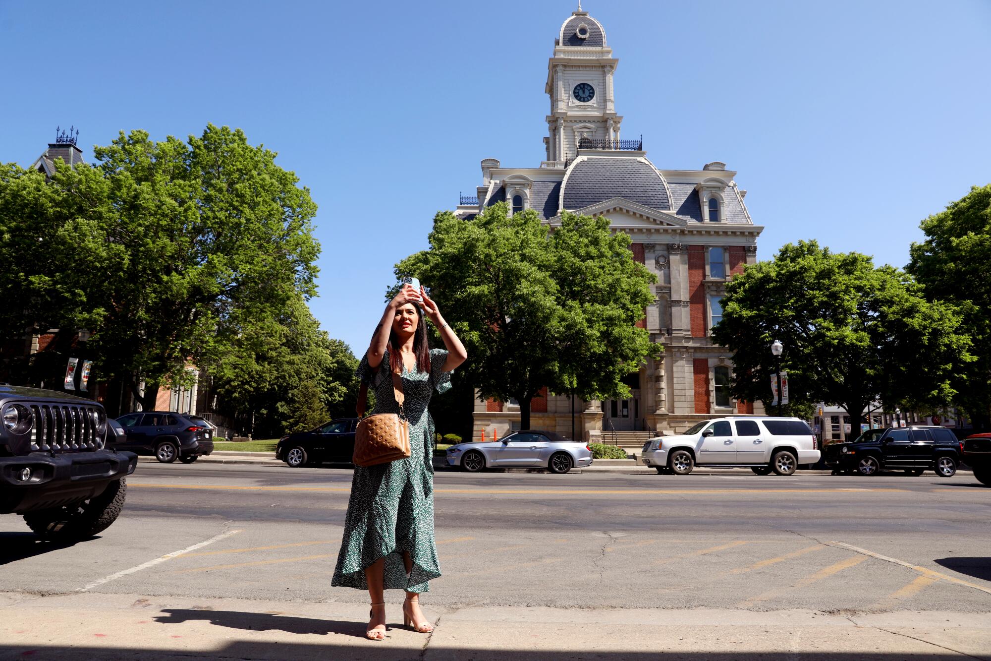 A woman stands on a sidewalk in front of a courthouse holding her phone up in the air