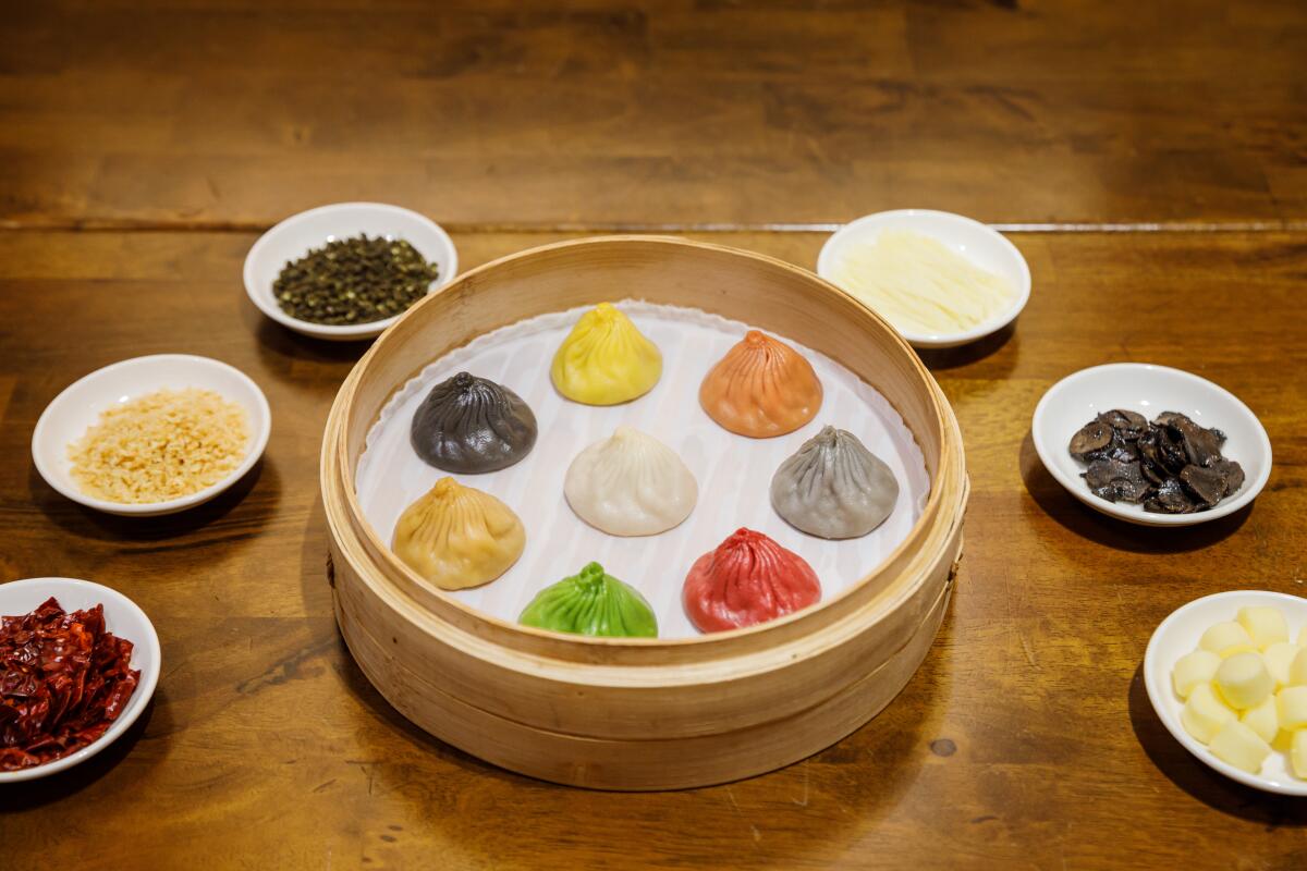 Eight xiao long bao in a round bamboo steamer, with each color representing a different flavor.