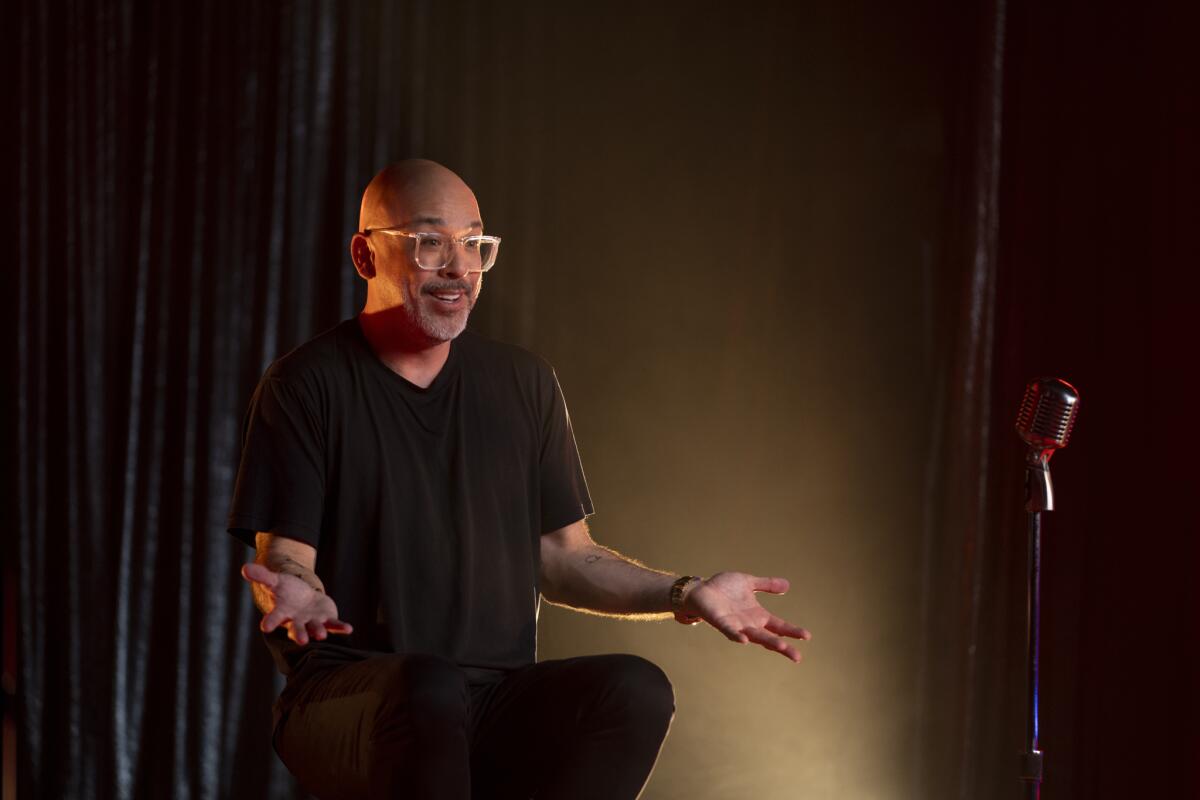 A man in clear-rimmed classes holds open his arms and smiles while sitting on a stool
