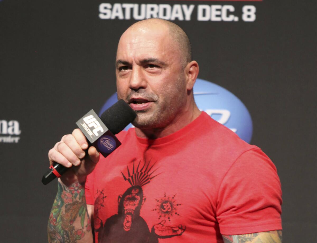 A bald, muscular man wearing a red shirt holds a microphone