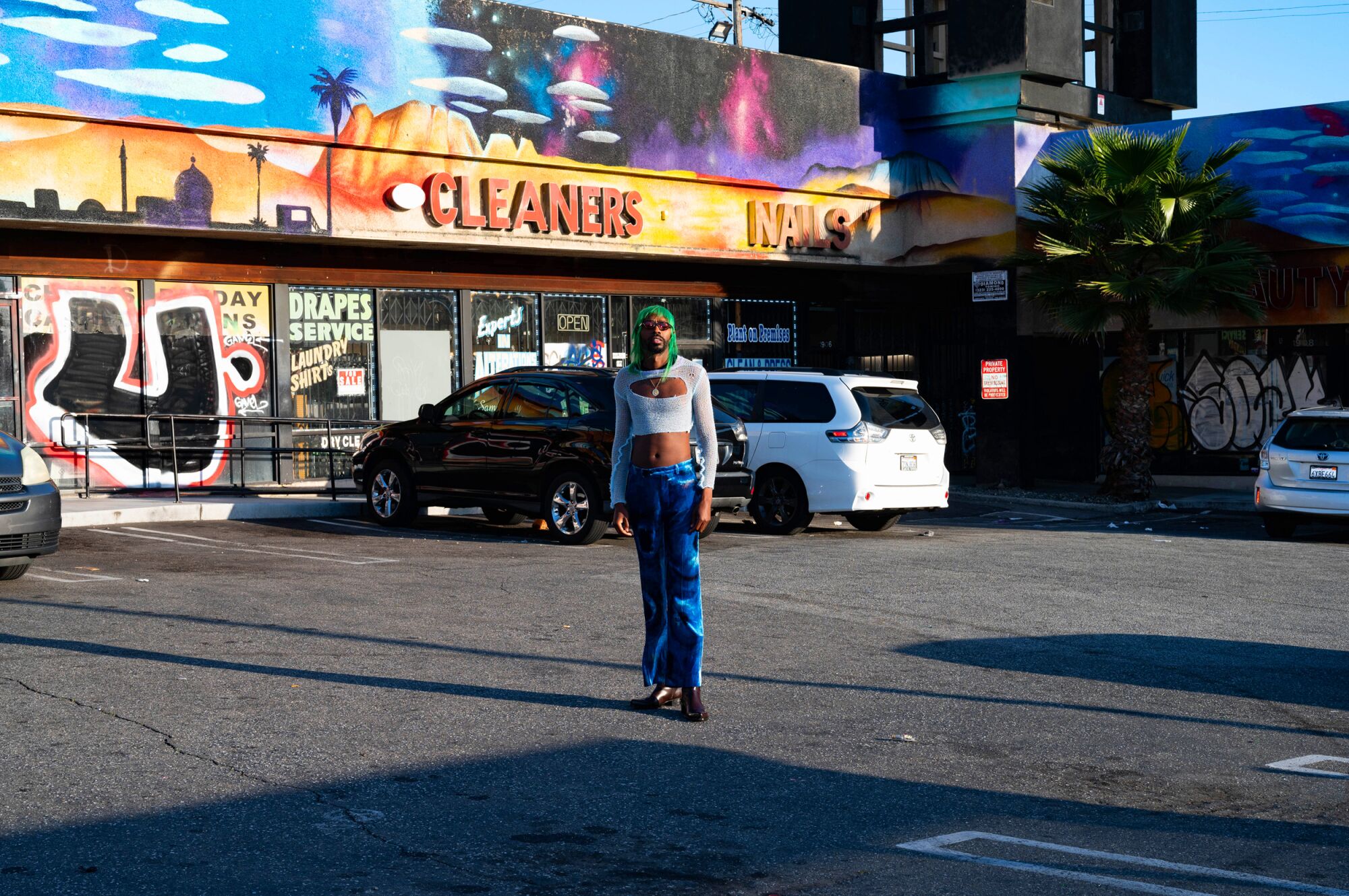 A person stands in the parking lot of a strip mall, with colorful signs reading "Cleaners" and "Nails" in the background.