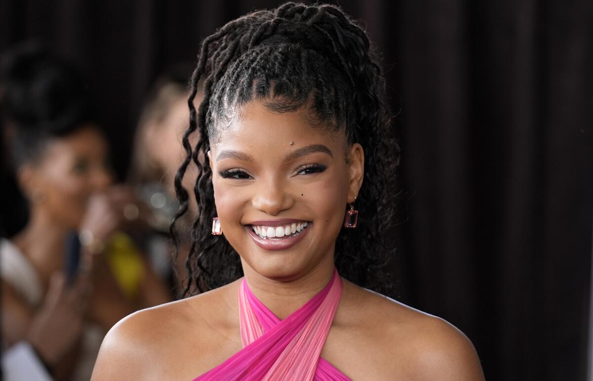 Halle Bailey in a pink halter dress with her hair in a high ponytail, smiling an posing against a black background