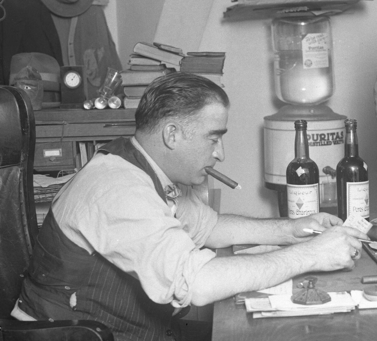 A man sits at a desk smoking a large cigar. Near him are two bottles of alcohol.