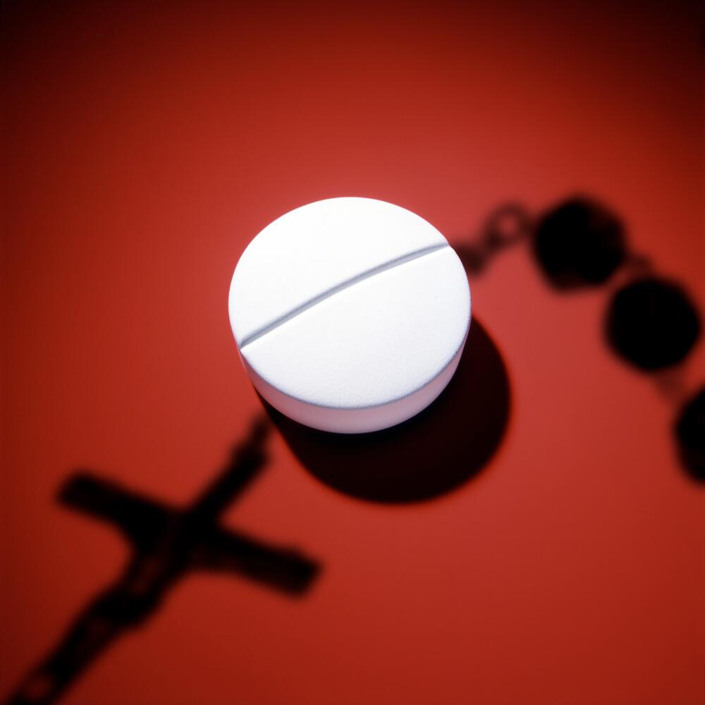 Photo illustration of a white pill on a red surface with a shadow of rosary beads cast across the pill.