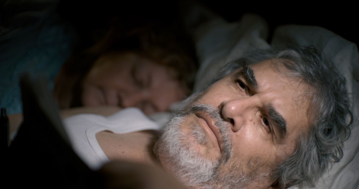 An older man awake in bed as his wife sleeps beside him.