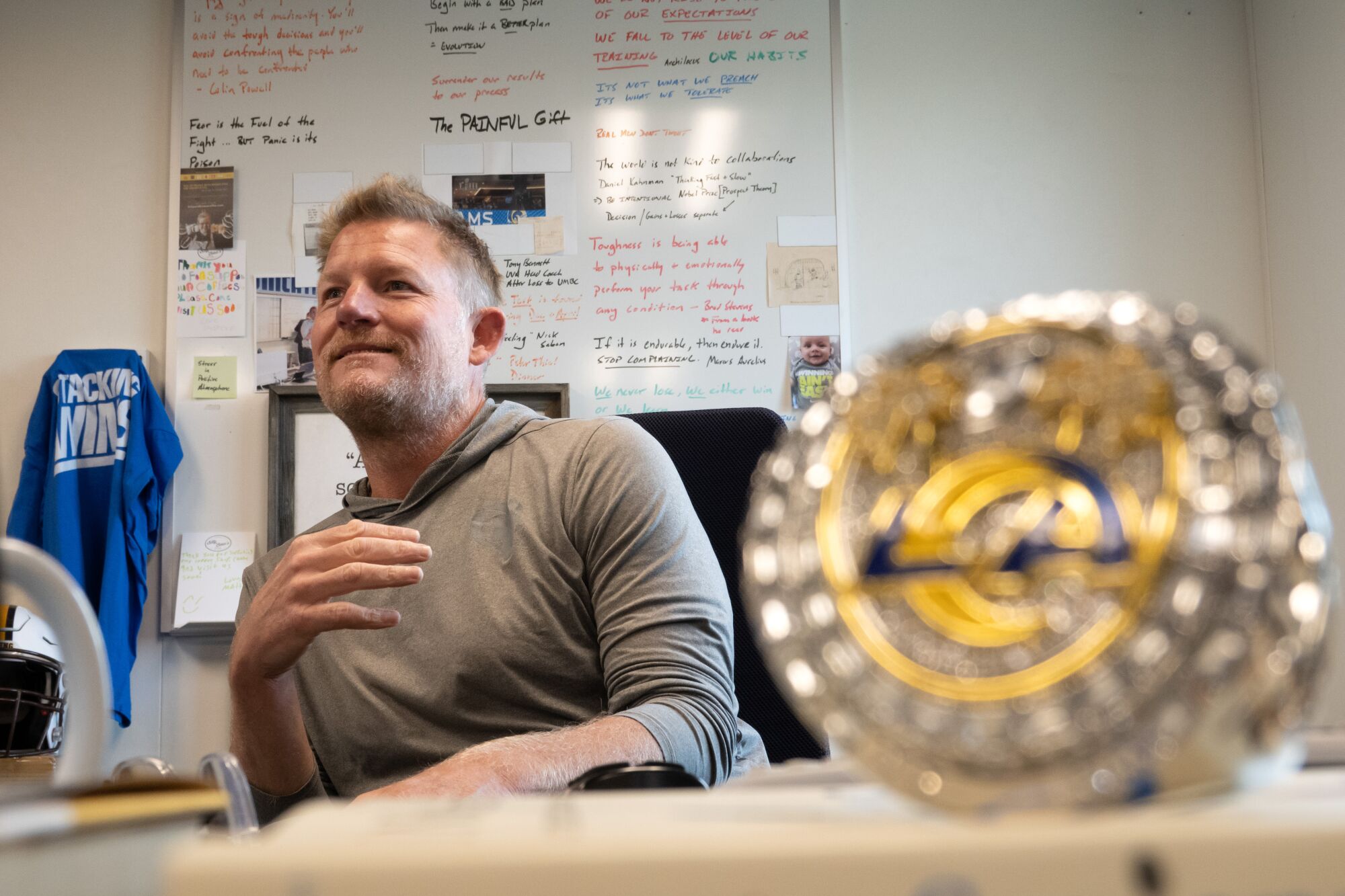 A man in a gray shirt sits in a black chair in front of a white board with a Rams Super Bowl ring in front of him.