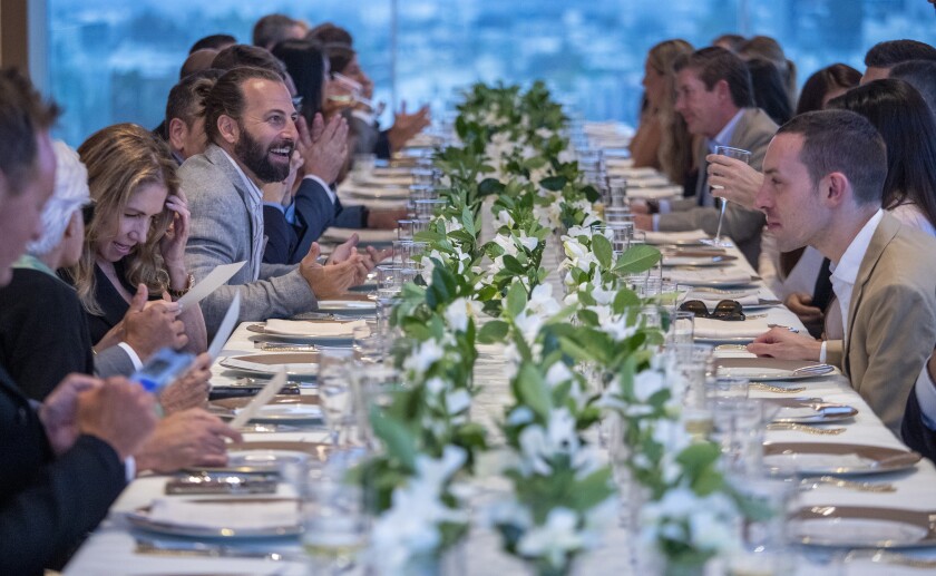 People, chatting, line either side of a long dining table with place settings and flowers.