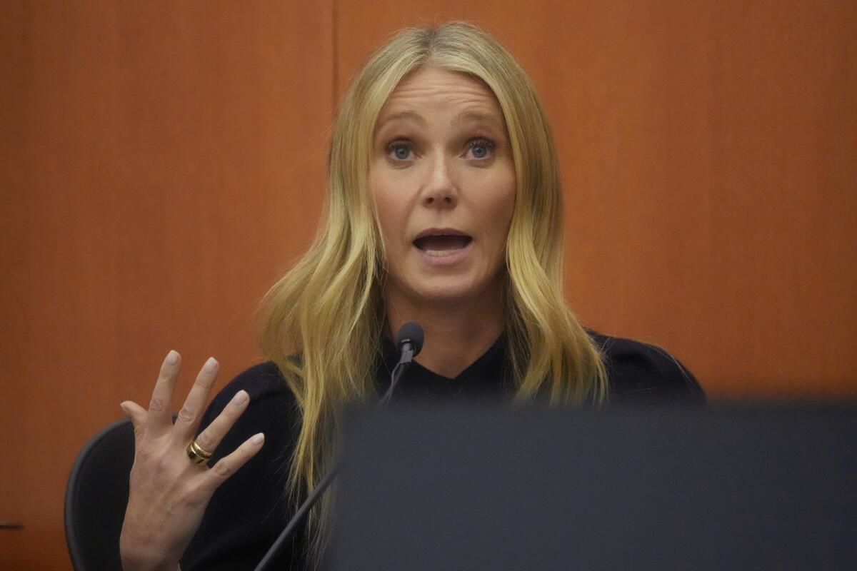 Gwyneth Paltrow, in a black shirt, gestures with one hand while speaking into a microphone on the witness stand.