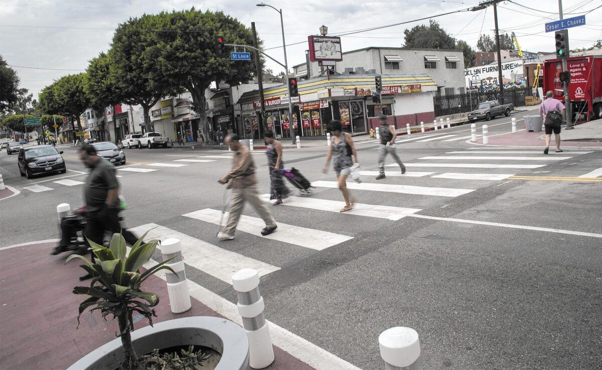 Pedestrians cross the street at Cesar Chavez Avenue and St. Louis Street. In an effort to slow traffic, the curbs have been extended and white bollards installed.
