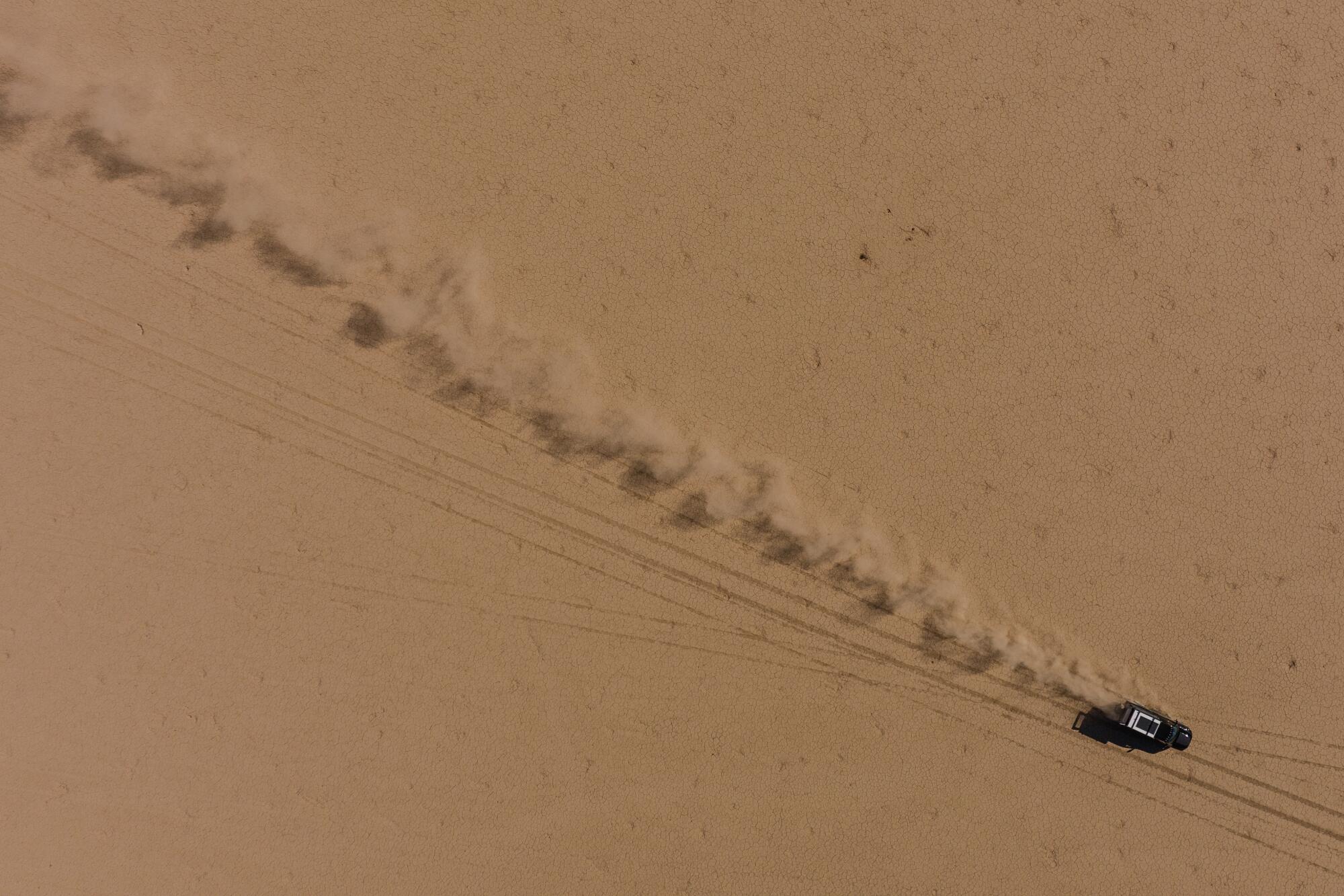 A drone photo of a truck driving across a dry lake bed, kicking up a stream of dust in its wake.