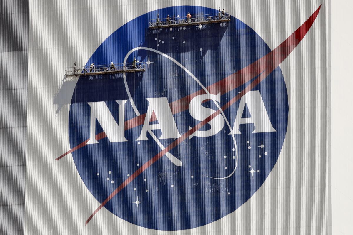 Workers on platforms suspended in front of a giant NASA logo
