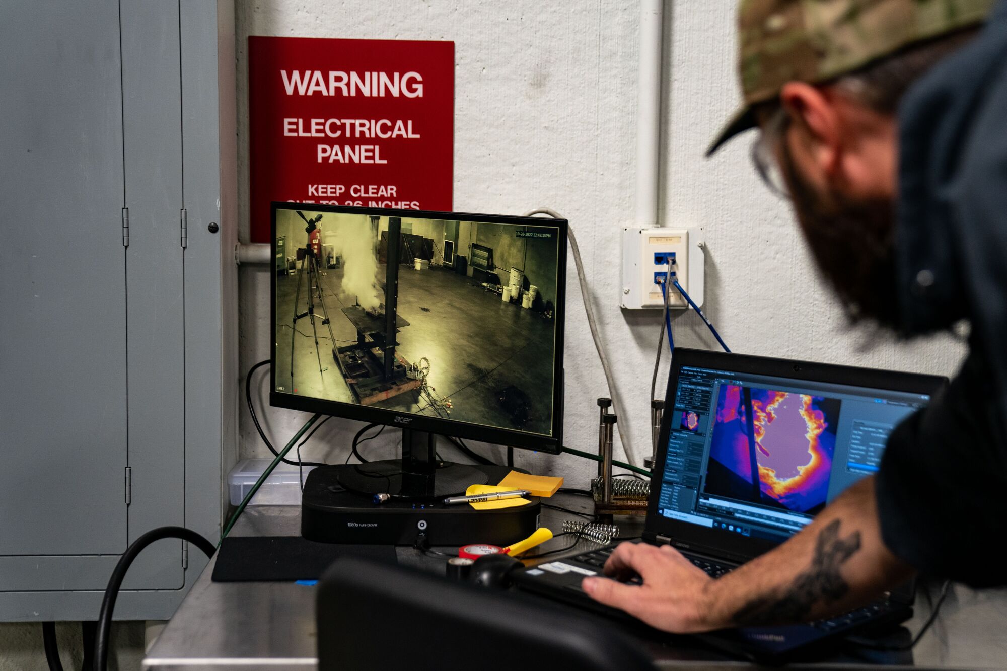 A bearded man wearing glasses and a camouflage cap, right, looks at two computer screens 