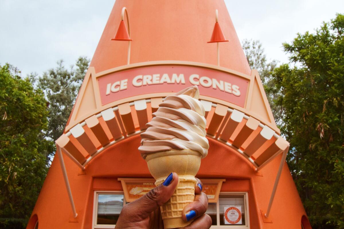 A hand holds up a chocolate and vanilla swirl soft-serve cone in front of an orange traffic-cone-shaped ice cream stand.