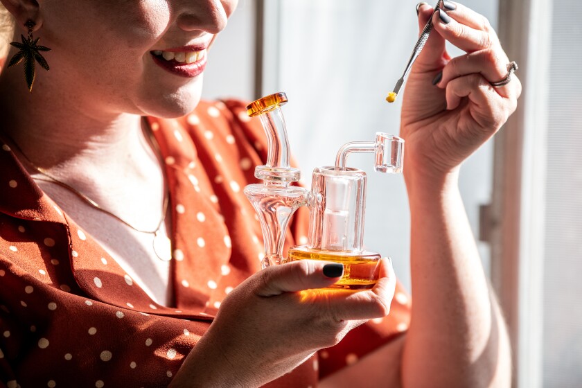 A woman holds a glass water pipe in one hand and a dab of THC concentrate in the other.