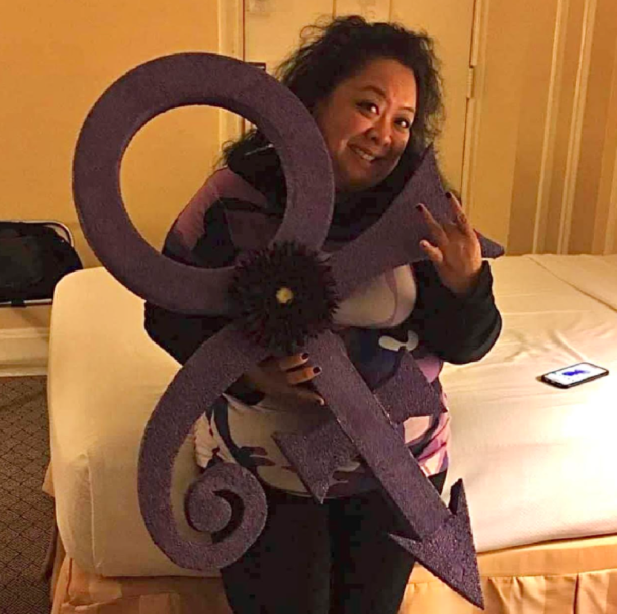 Woman poses with Prince Purple Love Symbol sign on hotel room bed. 