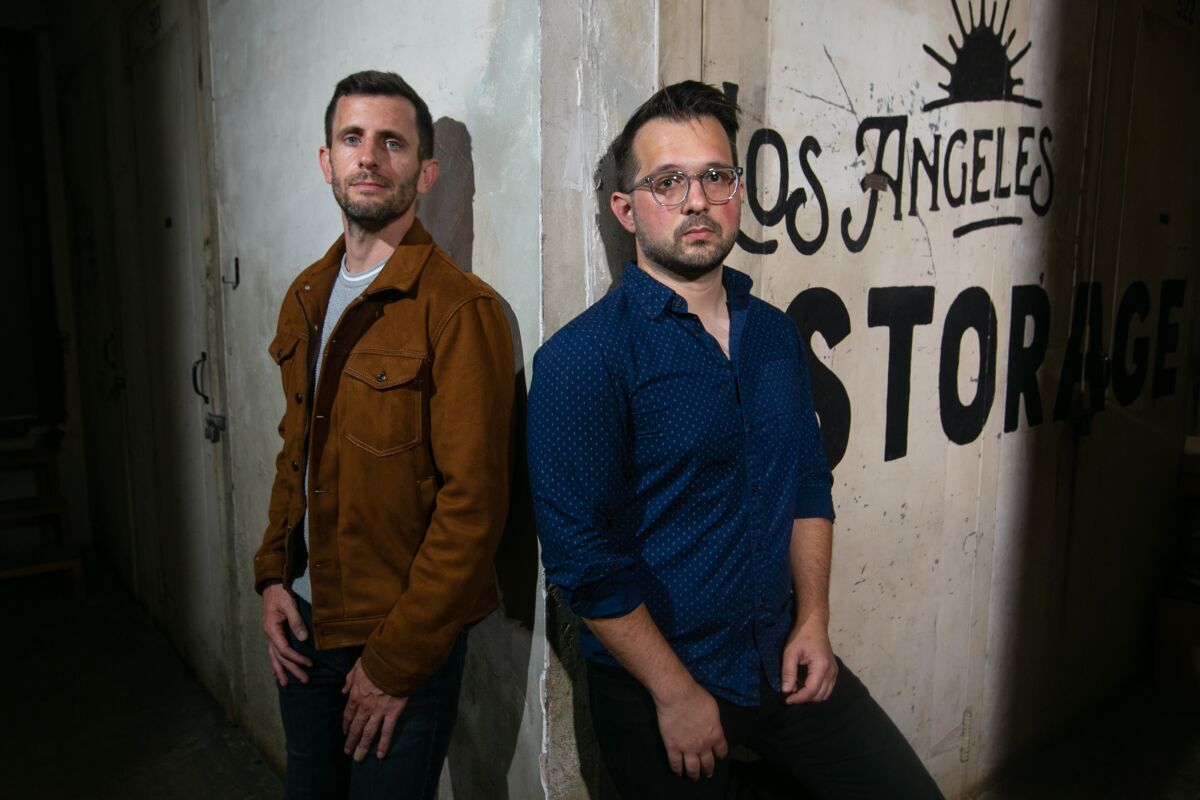 Two men lean against a wall with lettering that reads "Los Angeles Storage"