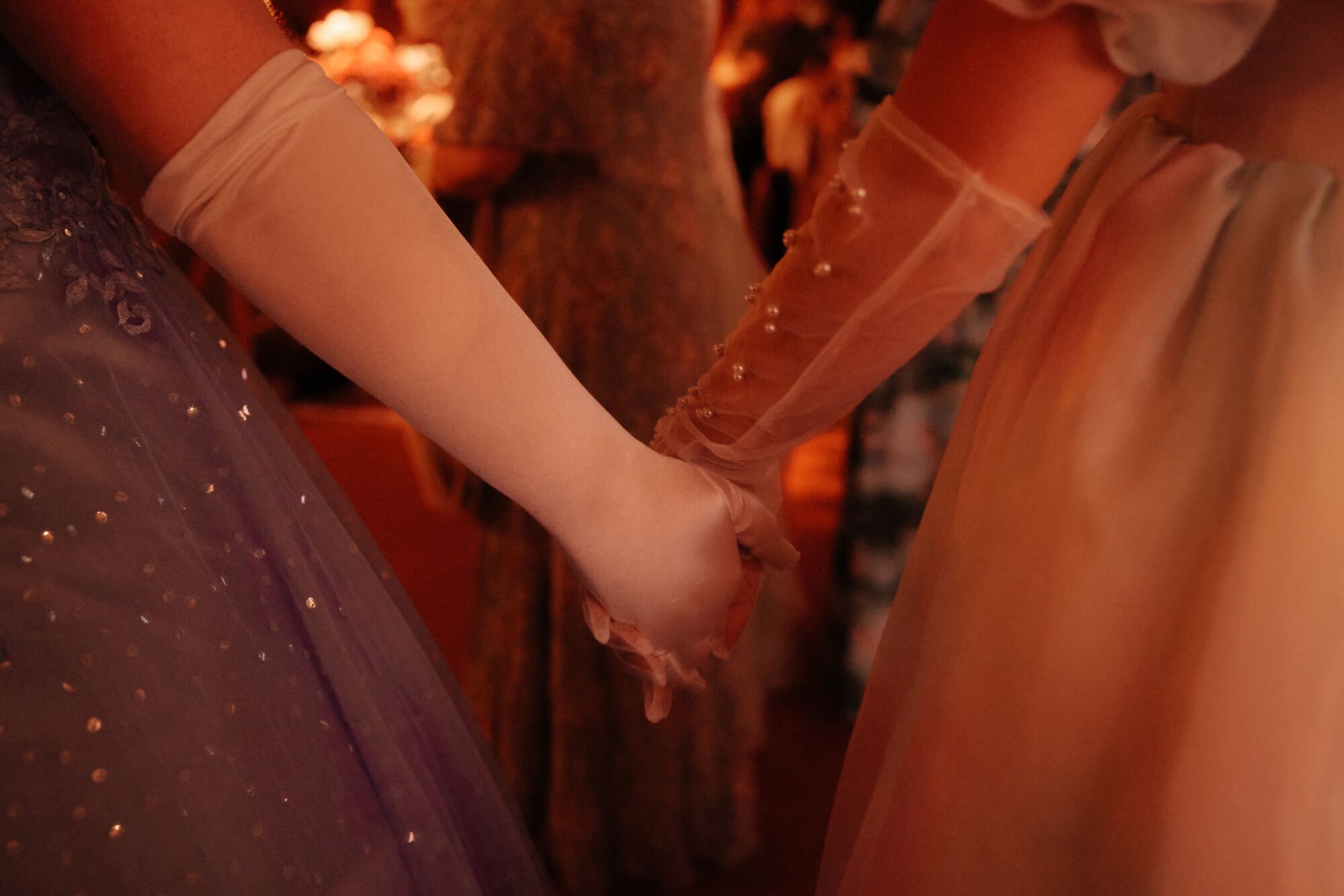 Two women in long gloves hold hands at a dress ball.
