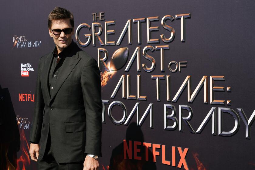Tom Brady wears shades while standing in front of a sign reading "The Greatest Roast of All Time: Tom Brady" 