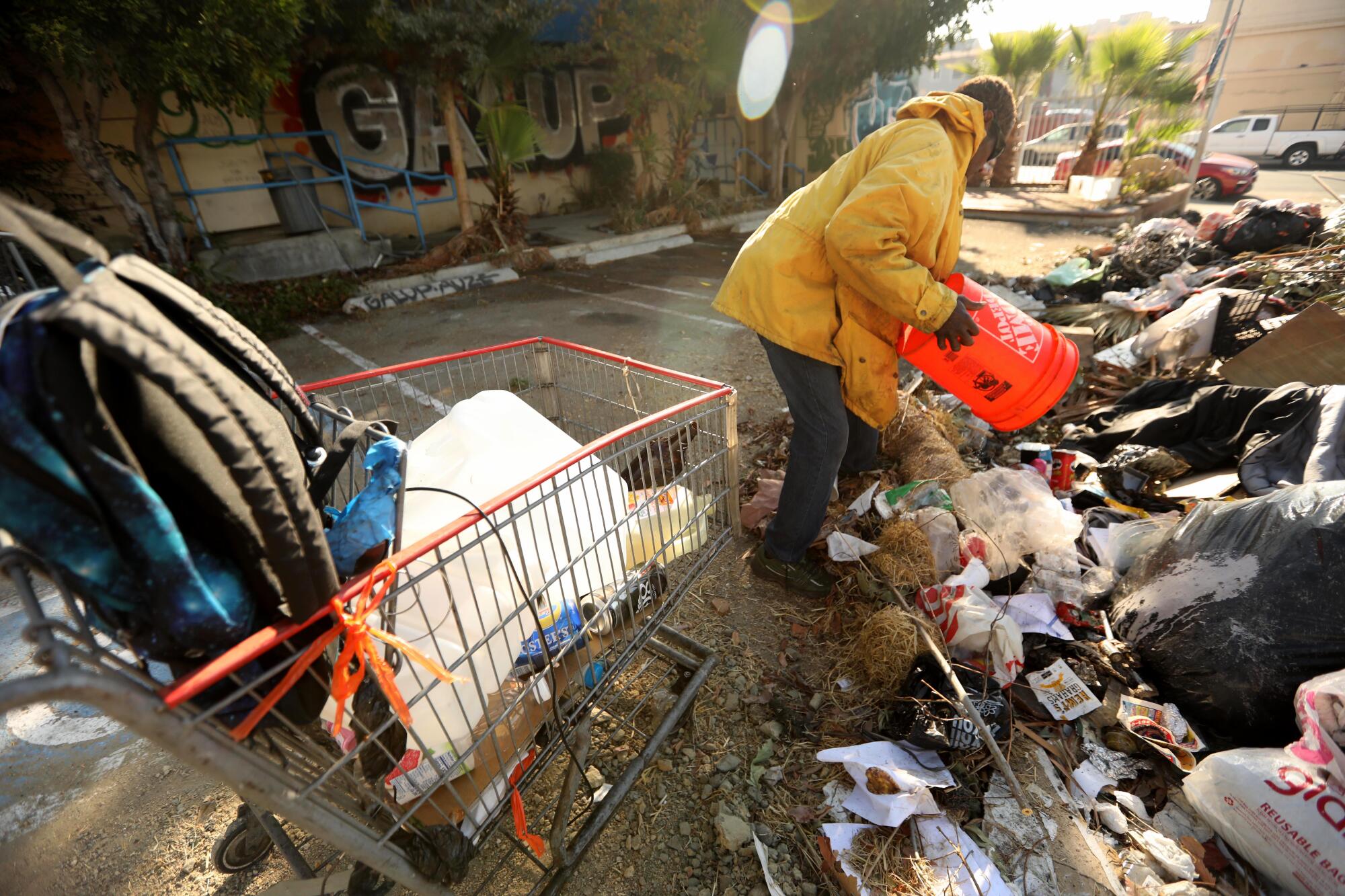 A man dumps an orange bucket of items onto a trash heap in a dirt lot