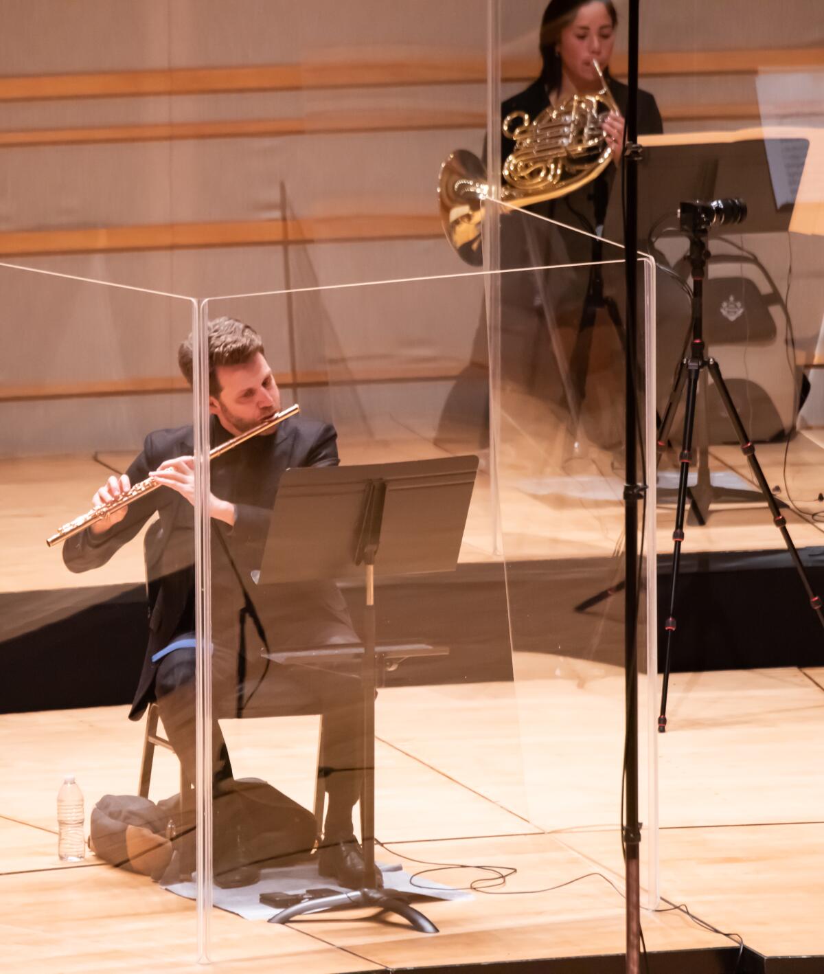 The brass and wind musicians of the Pacific Symphony perform encased by plexiglass.