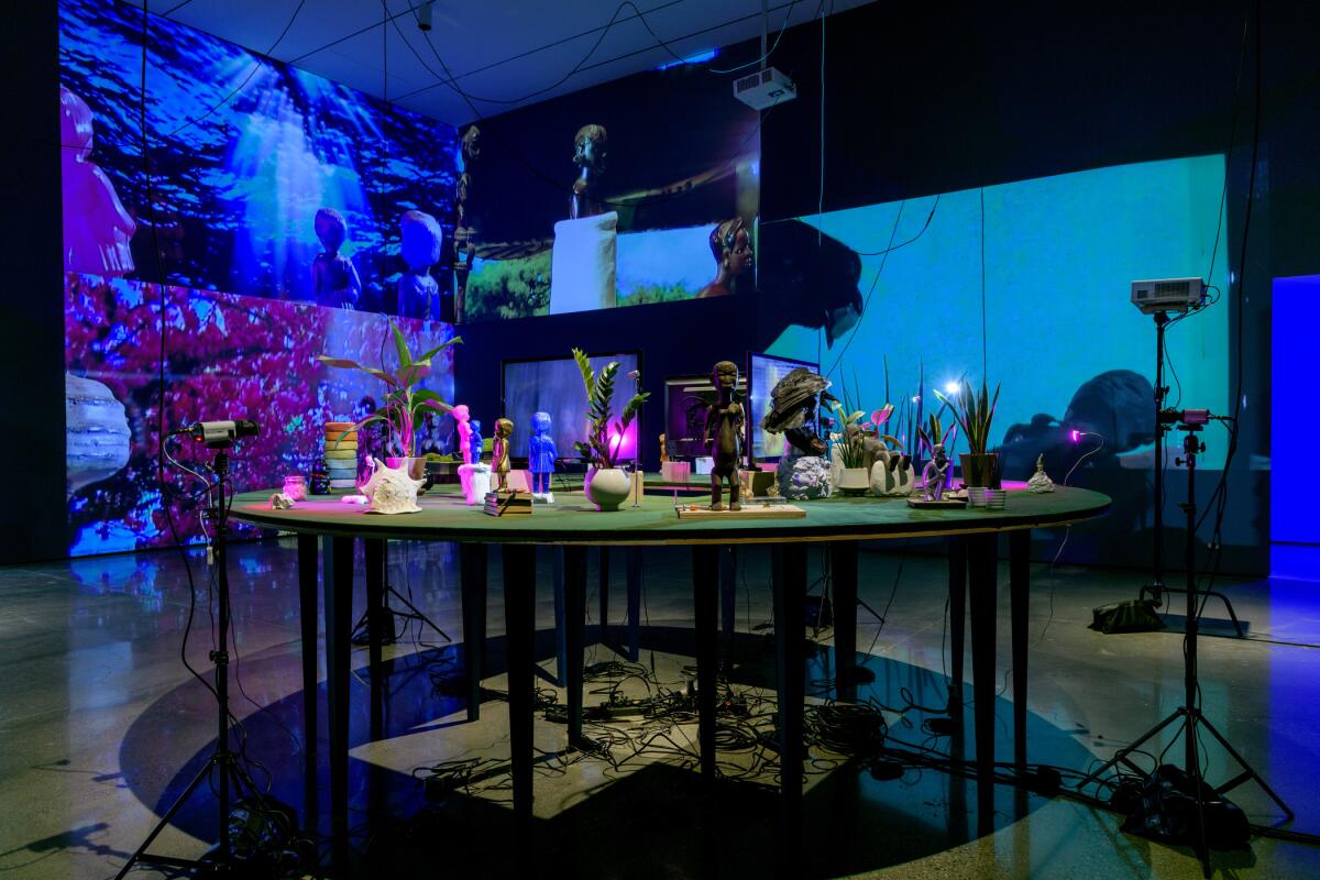 Statues, dolls and plants sit on a table under images projected on several surrounding walls.