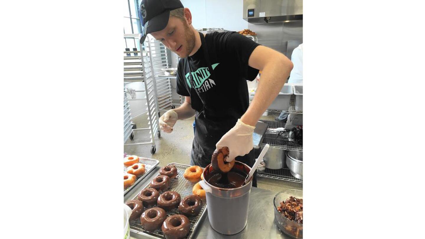 A worker at D'arts Donut Shop hand-dips doughnuts in a bucket of chocolate.