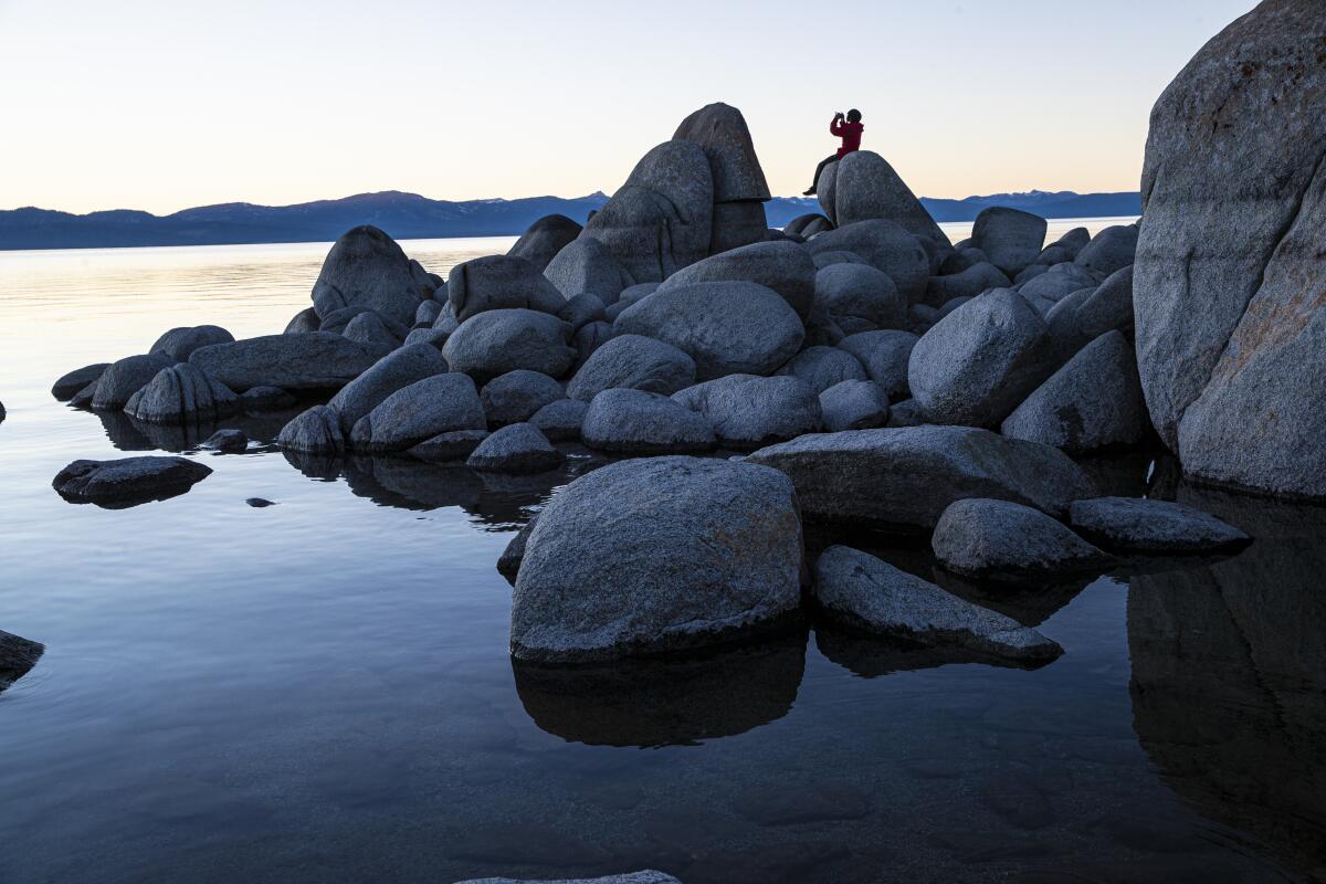 A person sits on a large rock among other big rocks overlooking water.