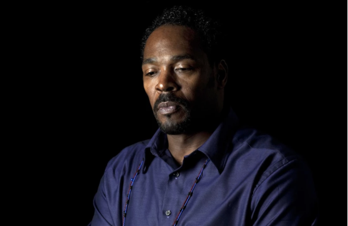 A photo portrait of a thoughtful-looking man in a blue shirt against a dark background.