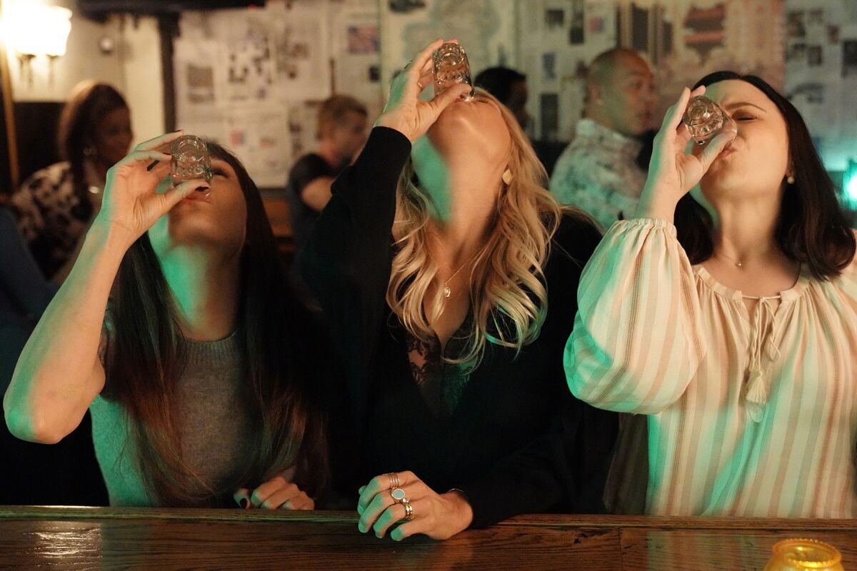 Three women at a bar tilt back their heads as they down shots of liquor.