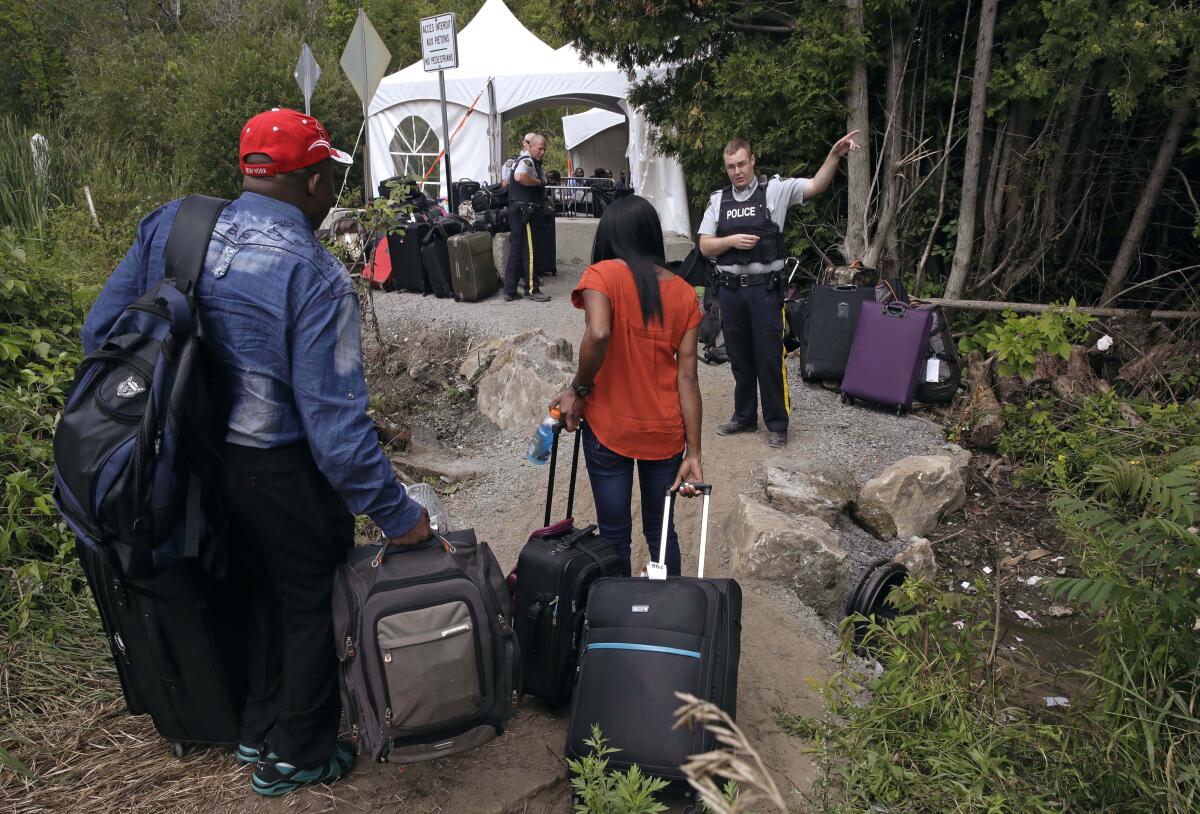 A police officer directs migrants walking a dirt path with suitcases.