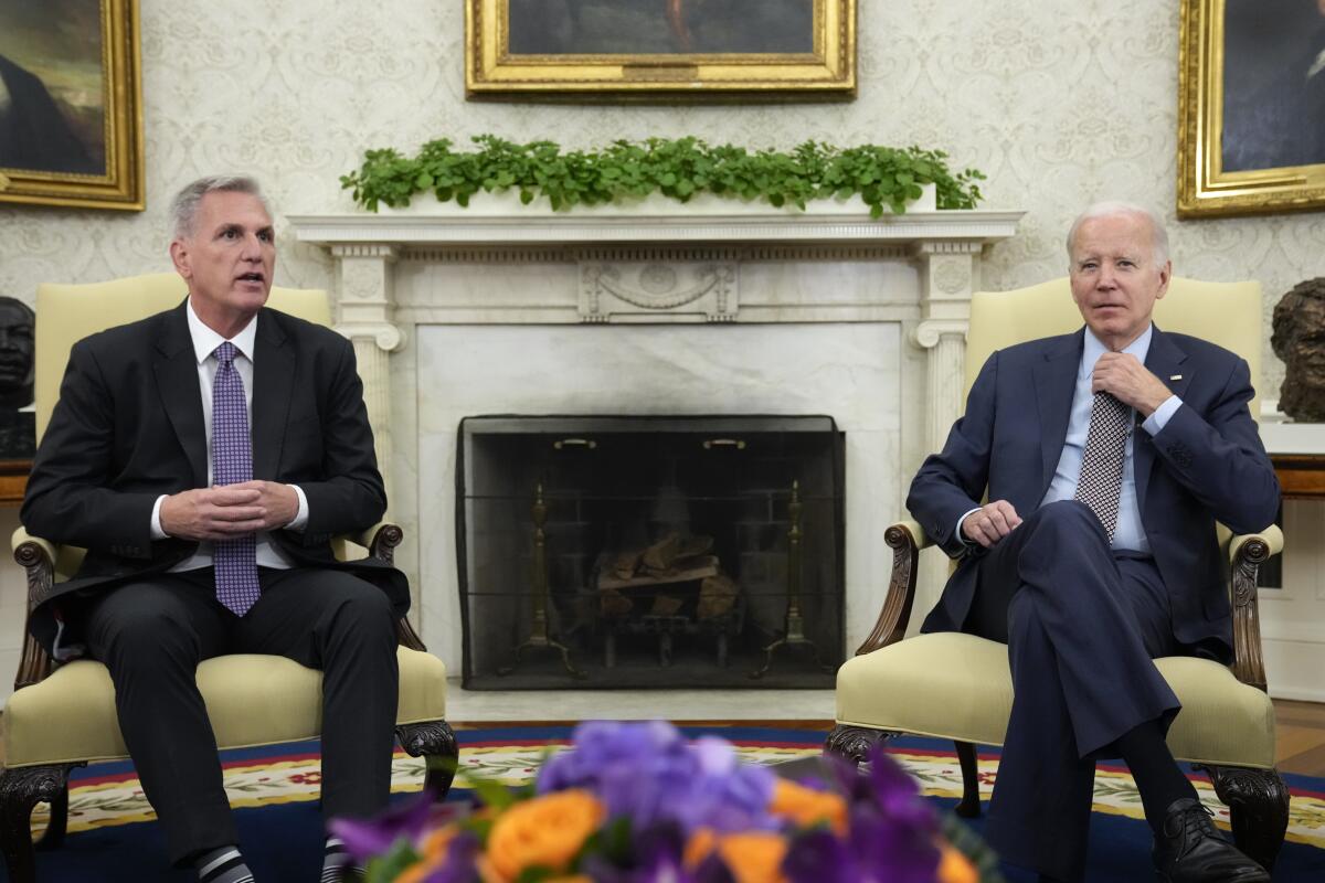 House Speaker Kevin McCarthy and President Biden, both wearing dark suits, are seated in armchairs flanking a fireplace.