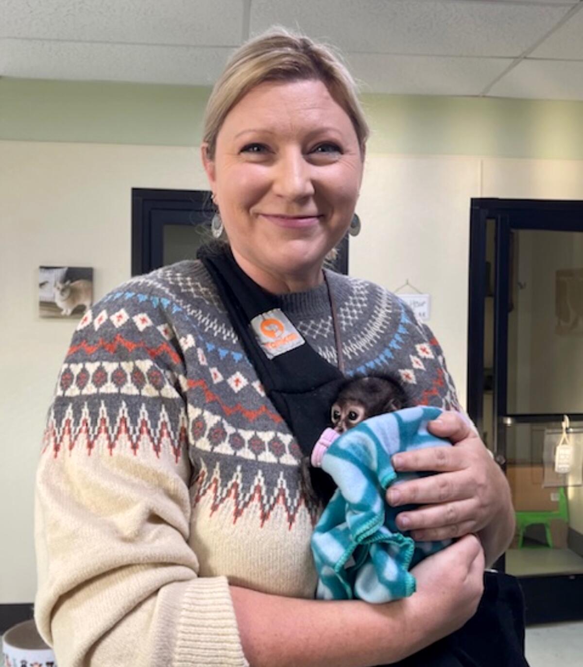 A smiling woman holds a tiny monkey wrapped in a blanket against her chest.