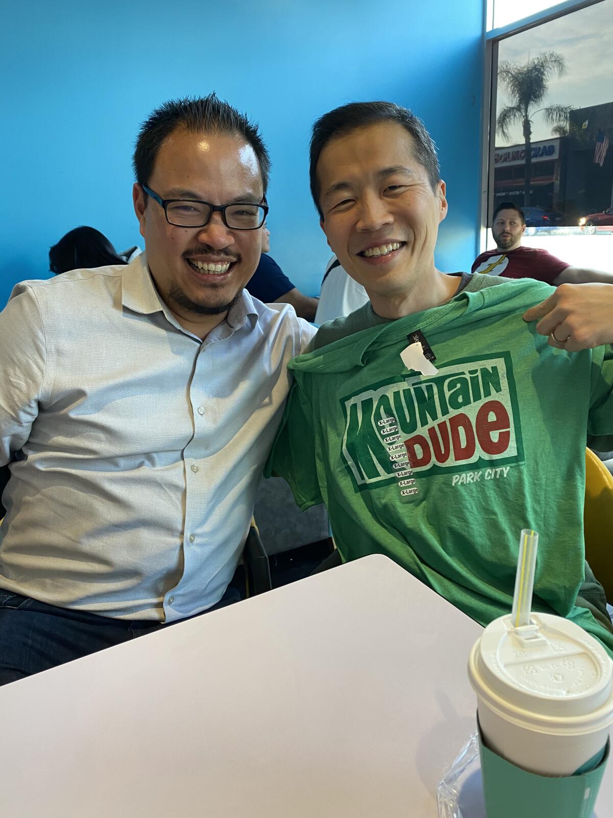 L.A. Times critic Justin Chang and Lee Isaac Chung at a table. Chung holds up a Mountain Dude T-shirt.