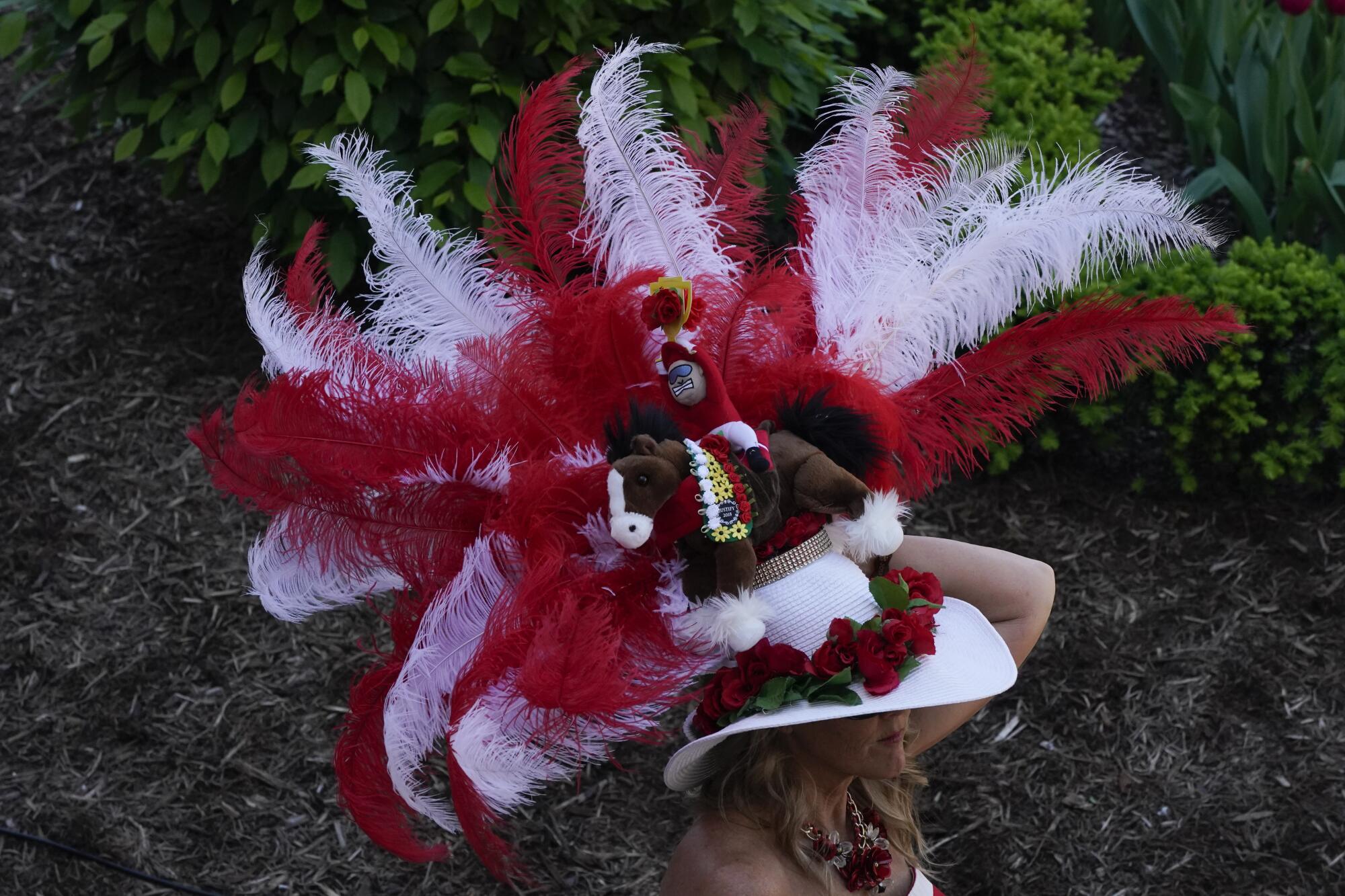 A women walks in a white brimmed hat with red roses around it and red and white feathers and a toy horse and rider above it.
