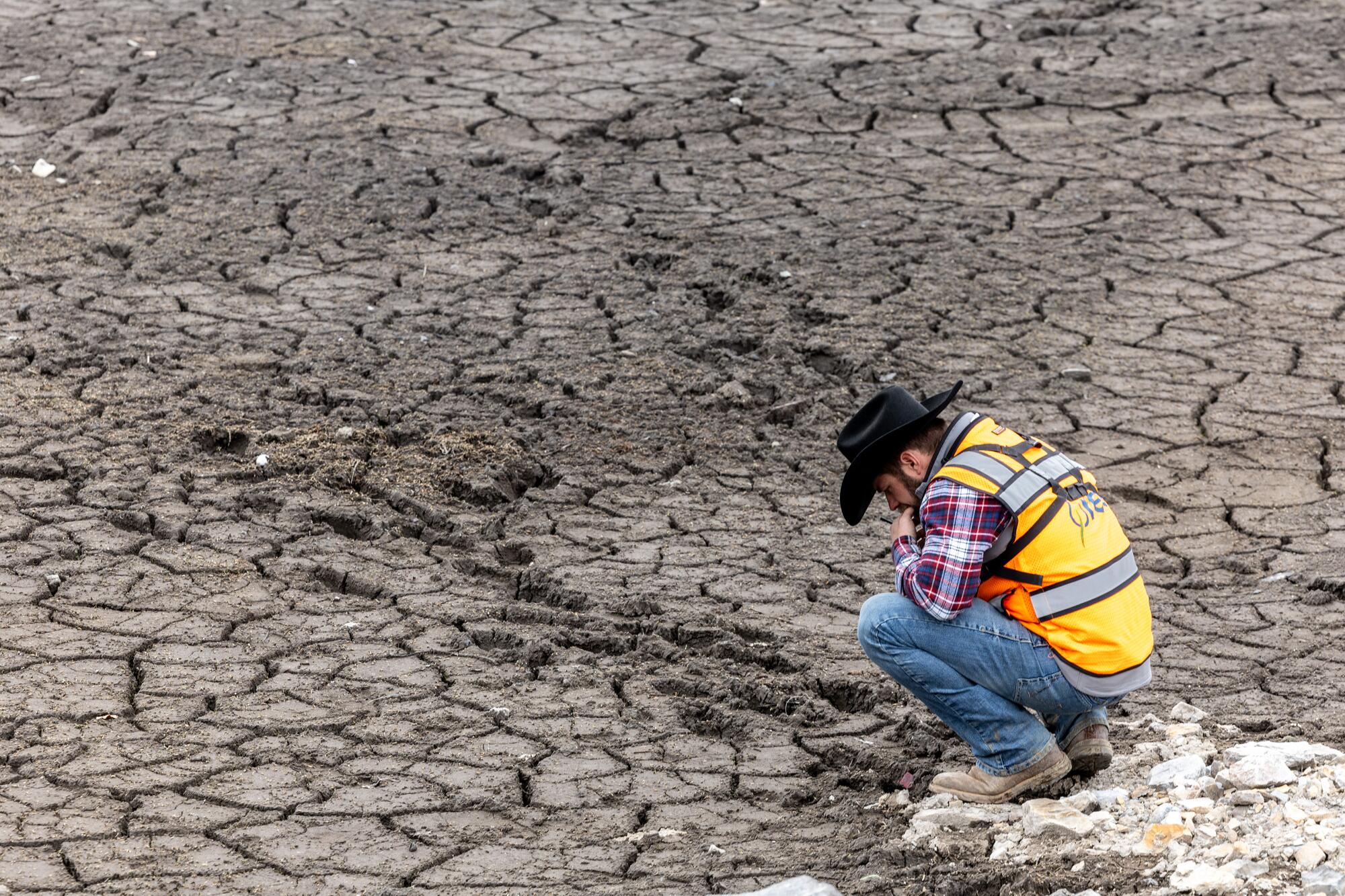 A man in a yellow safety vest squats on a dry spot next to footprints in an expanse of cracked mud, looking down