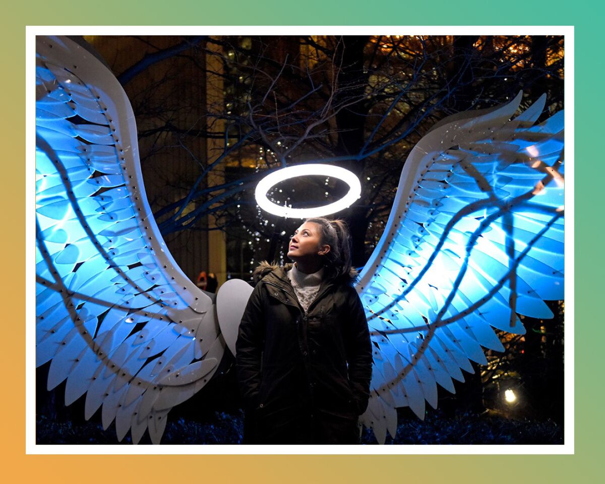 After dark, a woman stands with a sculpture of angel's wings behind her and a halo above.