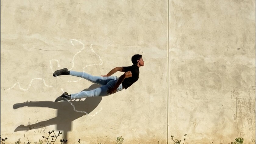 A dancer in a black shirt and blue tights sails in front of an ivory-colored wall with animated markings around her