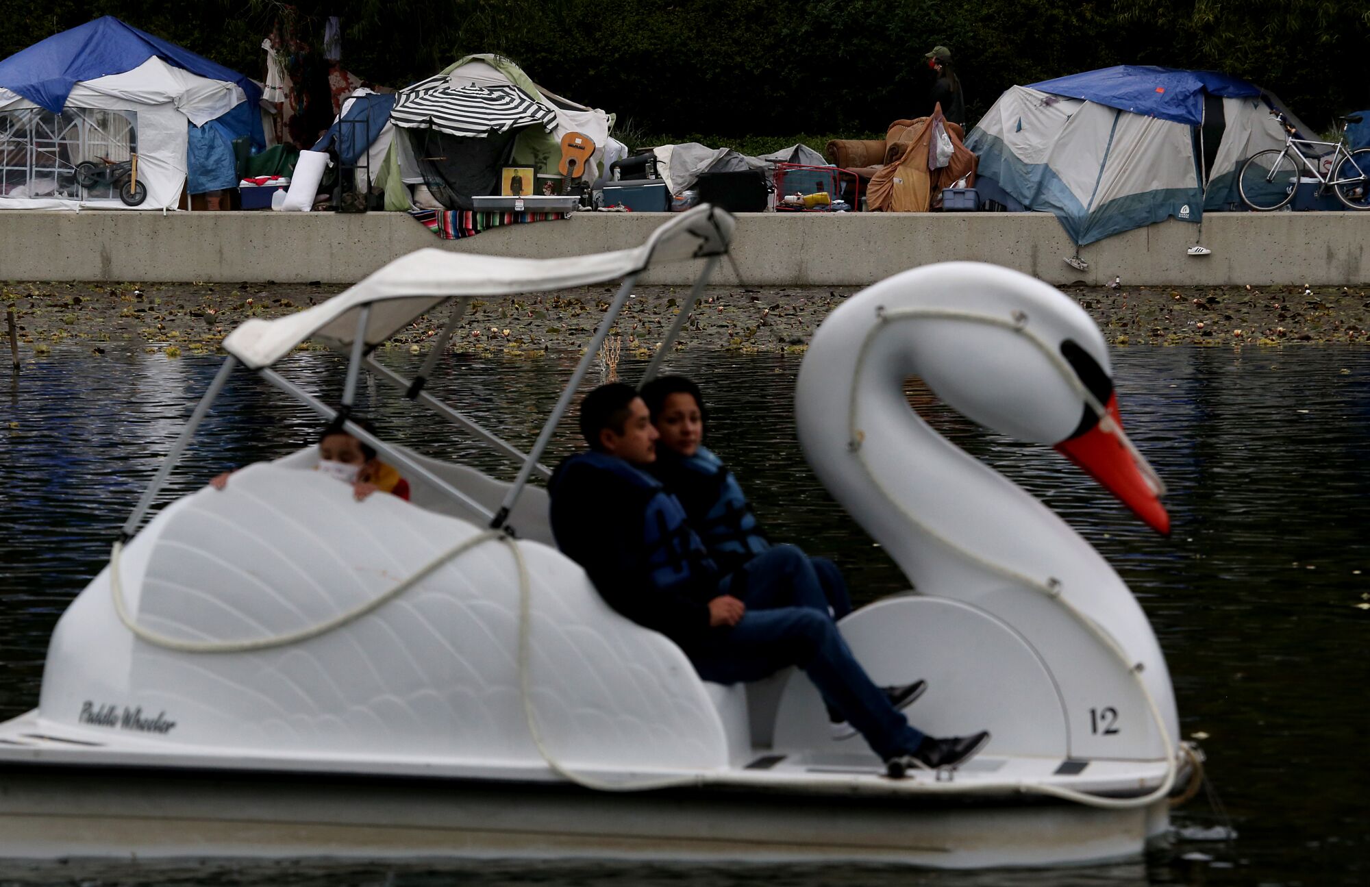 A couple use a swan boat at Echo Park Lake with a homeless encampment in the background.