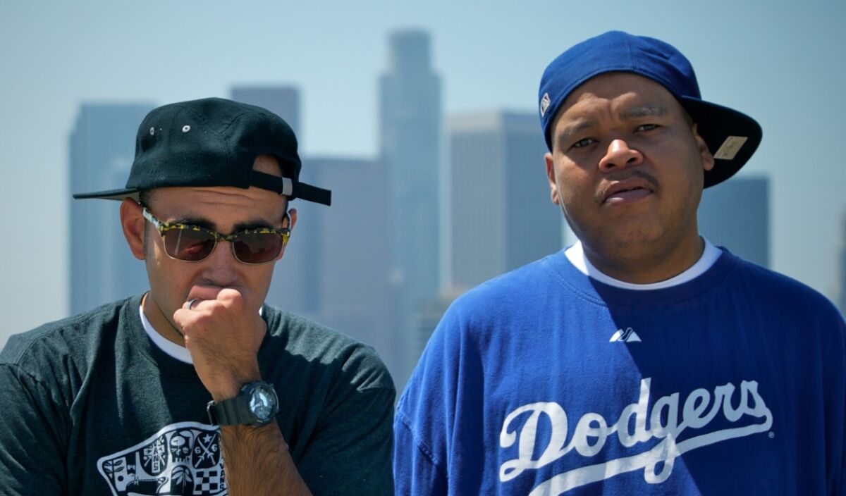 Two men in T-shirts and baseball caps pose side-by-side outdoors.