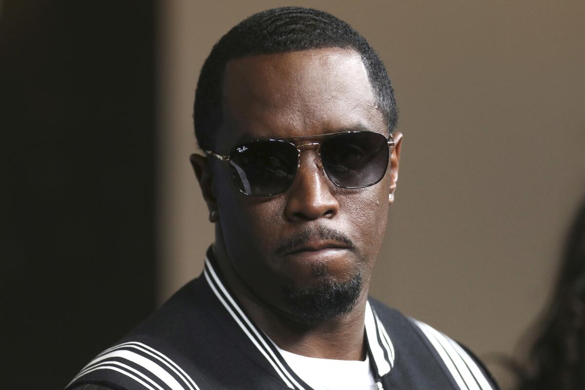 Sean "Diddy" Combs wearing a dark jacket with white stripes and big sunglasses with a somber expression on his face