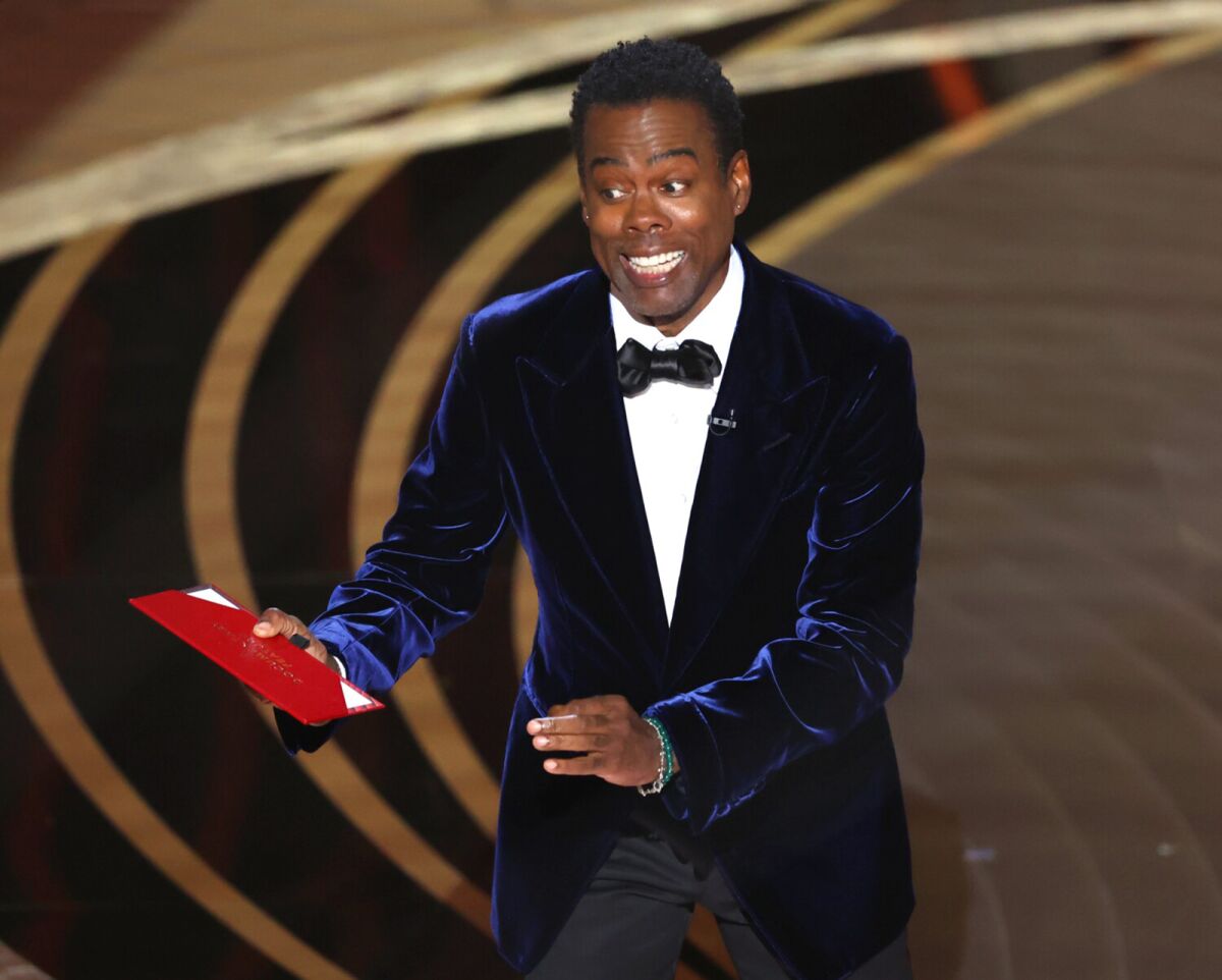 Chris Rock, holding a red envelope and wearing formal attire, speaks onstage during the Oscars