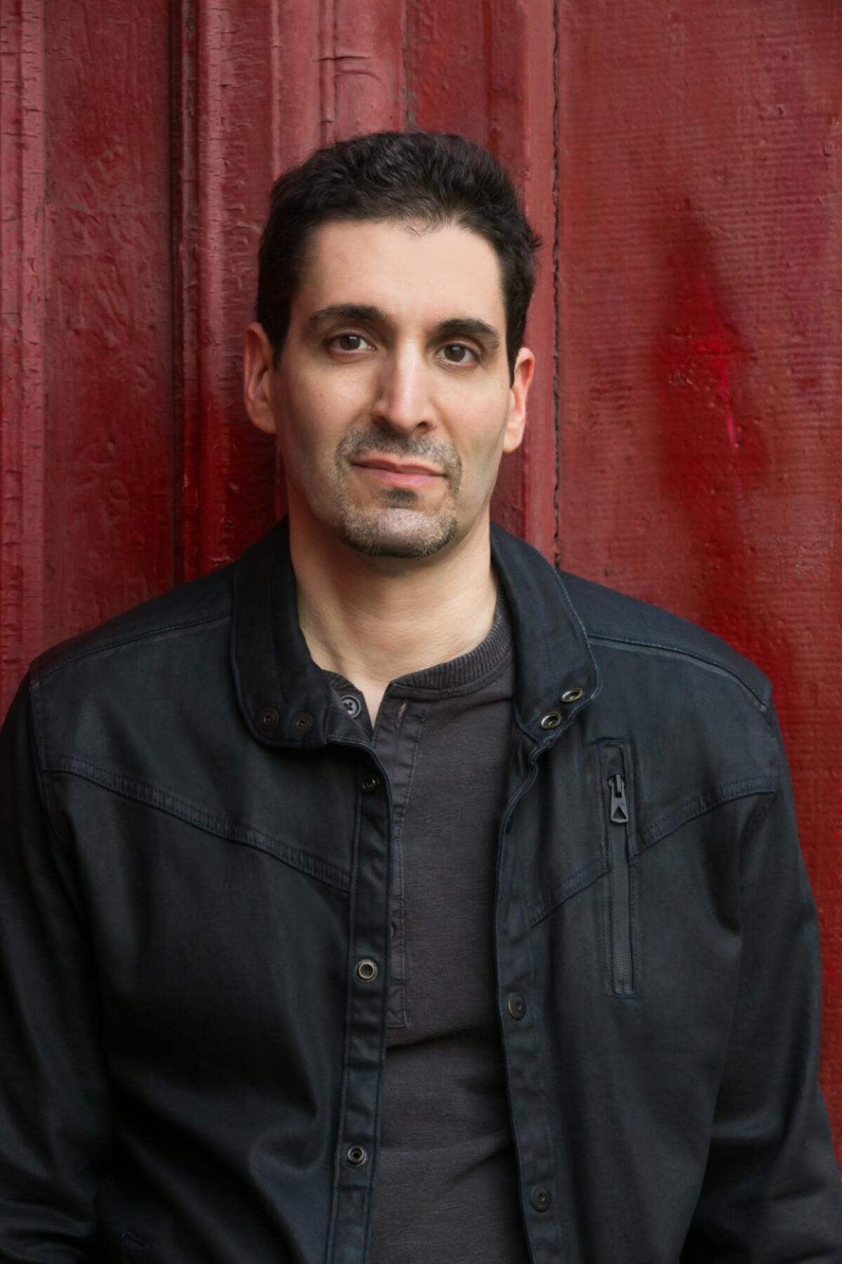 A portrait of a man in a black shirt and black jacket in front of a red curtain